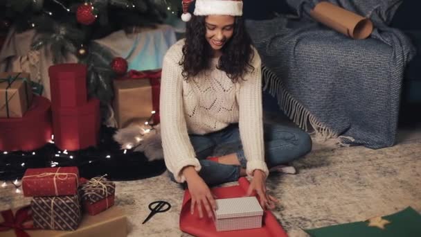 Atrakcyjna Mulatto dziewczyna w Mikołajach Hat pakowania prezenty siedzi pod choinką. Młoda Brunetka w białym pulowerze przygotowująca się do świąt. Koncepcja świętowania wakacji. — Wideo stockowe