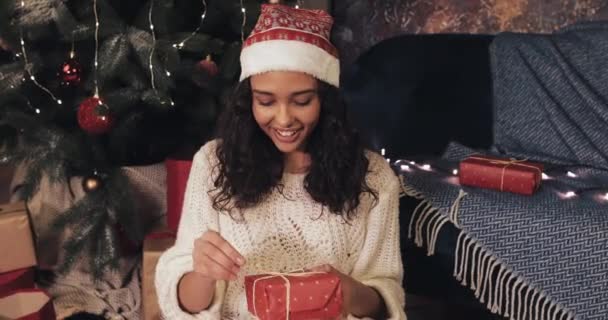 Krásná usmívající se Mulatto dívka sedí pod vánočním stromem na útulném domácím pozadí při rozbalování Red Present Box, Hledáte vzrušený. Koncept oslav Vánoc a Nového roku. — Stock video