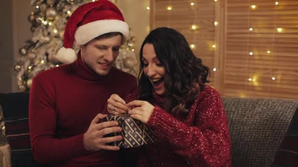 Junger glücklicher Mann mit Geschenkschachtel und Frauen, die eine Verbeugung darauf machen und aufgeregt auf dem Sofa in der Nähe des Weihnachtsbaums zu Hause sitzen Hintergrundkonzept von Feiertagen und Neujahr. — Stockvideo