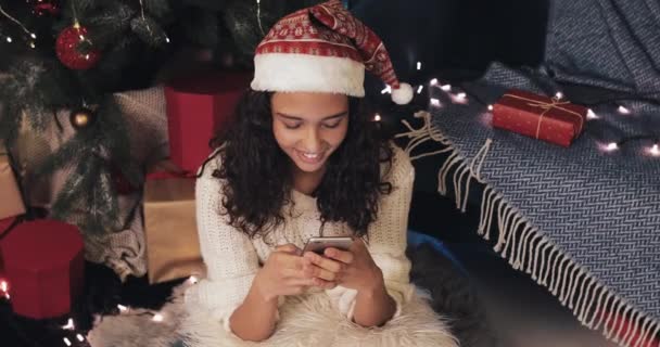 Attractive Brunette Mulatto Girl in Santas hat Greeting Friends with Christmas Using Smartphone Typing Messages Sitting at Floor near Christmas Tree. Communication and Holiday Concept. Top View — ストック動画