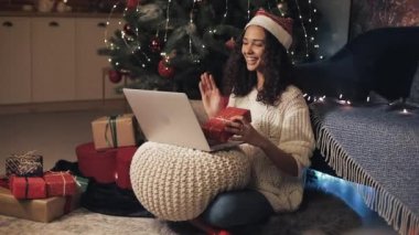 Noel Baba Şapkalı Genç Güzel Kadın 'ın dizüstü bilgisayarında, Noel Ağacı' nın yakınındaki Evinde Yerde Kameraya Hediye Kutusu Gösterirken görüntüsü var. Yeni Yıl ve Noel Kutlama Kavramı. Yan Görünüm.