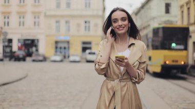 Akıllı telefonunu kullanarak kulaklık takan Güzel Genç Kız Müzik dinlemenin ve şehrin arka planında gülümsemenin tadını çıkarıyor..
