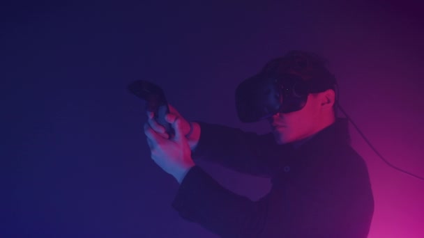 Joven hombre guapo en realidad virtual auriculares jugando juego de disparos en línea, el uso de controladores para disparar en el fondo de iluminación de neón cibernético. Primer plano . — Vídeo de stock