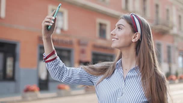 Mladý krásný kavkazský dívka s hnědými vlasy a čelenka na to pomocí svého smartphonu dělat video zprávu a foukání polibek a usmívání stojící na City Street. — Stock video