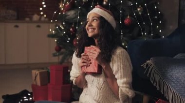 Noel Baba Şapkalı Genç Güzel Mulatto Kız süslenmiş Noel ağacının altında oturmuş, şimdiki kırmızı hediye kutusunu kucaklıyor, mutlu görünüyor. Noel ve Yeni Yıl Tatil Kutlaması Konsepti.