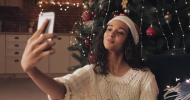 Genç Çekici Mulatto Kız Noel Şapkalı Süslü Noel Ağacı 'nın yanında oturuyor Samrtphone' uyla selfie çekiyor ve gönderiyor. Tatil ve Yeni Yıl kavramı.