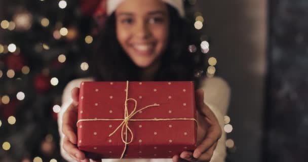 Close-up van Happy Beautiful Mulatto Girl in Santas Hoed Het aanbieden van geschenkdoos, Op zoek naar camera en glimlachen Staande op decoratieve verlichting achtergrond. Mensen en vieringen Concept. — Stockvideo