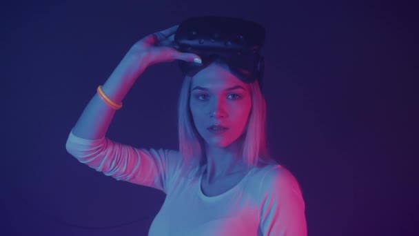 若い魅力的な白人ブロンドの女の子の肖像頭の上に仮想現実の眼鏡を保持します。,カメラを探して,ネオンの照明色に立って抽象的な背景ズーム. — ストック動画