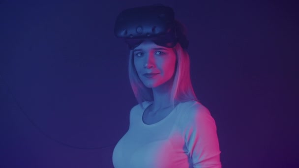 Retrato de una joven rubia atractiva con auriculares de realidad virtual, con suéter blanco mirando a la cámara, de pie de lado en colores de iluminación de neón Fondo abstracto . — Vídeos de Stock