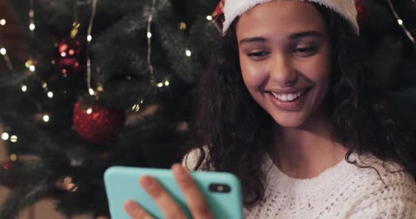 산토스 모자를 쓰고 크리스마스 트리에 앉아 스마트폰을 사용하며 스크린을 보고 웃으며 행복해 하는 아름다운 mulatto 소녀. 명절 컨셉이야. 가까이 서 — 비디오