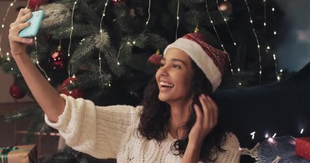 Junges attraktives Mulattin mit Christmütze sitzt neben geschmücktem Weihnachtsbaum, macht Selfie, posiert, zeigt Zunge, lächelt. Konzept von Feiertagen und Neujahr. — Stockvideo