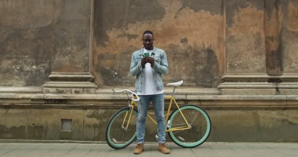 Tiro de cuerpo completo de hombre afroamericano con auriculares usando su teléfono inteligente y sonriendo mientras está parado frente a la bicicleta moderna y la pared del edificio antiguo en el fondo . — Vídeo de stock