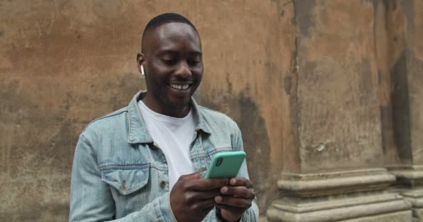 Gros plan sur un Afro-Américain dans Jean Jacket avec des écouteurs utilisant son smartphone et écoutant de la musique, regardant excité alors qu'il se tenait debout sur le vieux mur du bâtiment . — Video