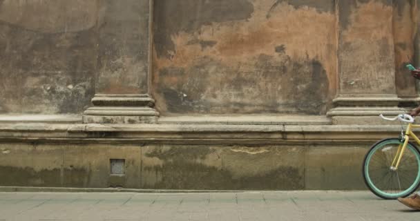 Афро-американский молодой человек в наушниках, используя свой современный мобильный телефон и толкая стильные тонкие шины велосипед во время прохождения по старой стены здания . — стоковое видео