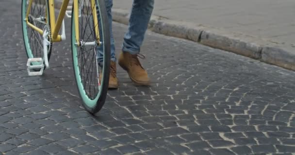 Primer plano de tiro de las piernas en pantalones vaqueros y botas casuales caminando en el viejo pavimento de piedra cerca de la bicicleta de neumáticos finos amarillos modernos. Vista lateral . — Vídeos de Stock