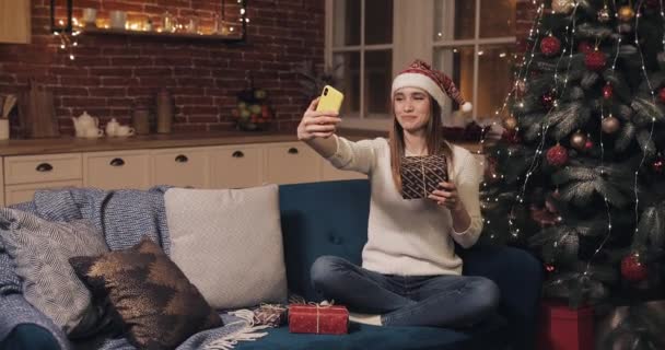 Leende kvinna bär Christamas hatt Sitter på soffan på Cosy Home Bakgrund håller och skakar presentförpackning nära julgranen gör ett videosamtal eller videomeddelande. Begreppet semester och nyår — Stockvideo