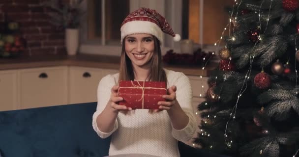 Portrét mladých kavkazských žen v klobouku Santas sedí na pohovce u vánočního stromku v útulné domácí vánoční pozadí dávat červený dárek. Koncept svátků a Nový rok. — Stock video