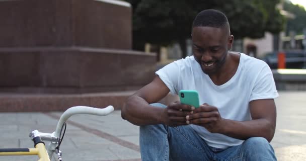 Közelkép Jóképű afro-amerikai srác alkalmi ruhák Használja a Smartphone, Izgatott, miközben ül a lépcsőkön a városban Bike a háttérben. Elülső nézet — Stock videók