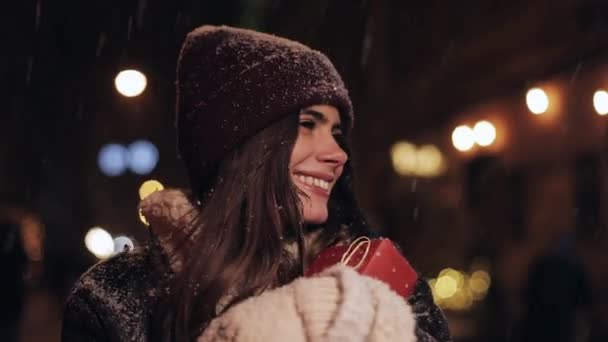 De cerca Retrato de Joven Chica Atractiva Feliz en Sombrero de Invierno y Mitones, Llevando Caja de regalo, Caminando en la caída de la nieve, Sonriendo, Mirando a un lado en las luces de la calle de la noche Fondo . — Vídeos de Stock