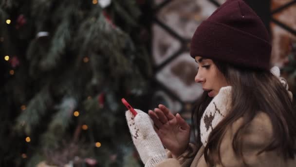 Закрыть молодой девушки носить зимнюю шляпу и варежки, стоя снаружи и используя свой смартфон, печатая, глядя удивительно, на фоне украшенного окна магазин. Вид сбоку . — стоковое видео
