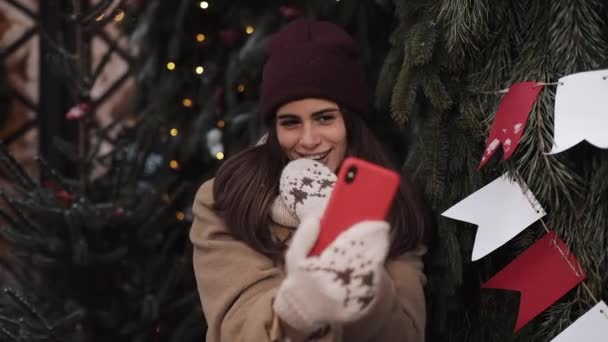 Kış şapkası, atkı, eldiven, poz verme ve gülümseme takan, selfie veya video mesajı çeken, Noel süslenmiş arka planda ayakta duran güzel kız. Kış Tatili Konsepti. — Stok video