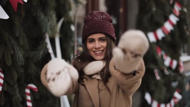 Усміхаючись молода щаслива дівчина в зимовому одязі стоїть в падаючих сніжинок зовні, посміхаючись, танцюючи, рухаючи руки, радіючи на Різдвяному прикрашеному тлі. Концепція зимового свята . — стокове відео