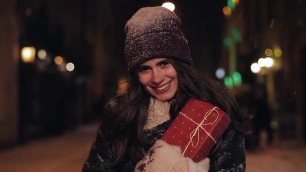 Portret młodej szczęśliwej atrakcyjnej dziewczyny w czapce zimowej i rękawiczkach, trzymającej obecne pudełko, chodzącej w opadającym śniegu, uśmiechniętej do kamery na tle świateł ulicznych wieczornych. Zamknij się.. — Wideo stockowe