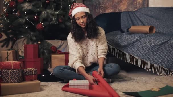 Krásná šťastná žena v Santas klobouk a bílý svetr připravuje dárky, řezy balicí papír, sedí pod vánoční strom na útulném domácím pozadí. Koncept svátkové oslavy. — Stock video