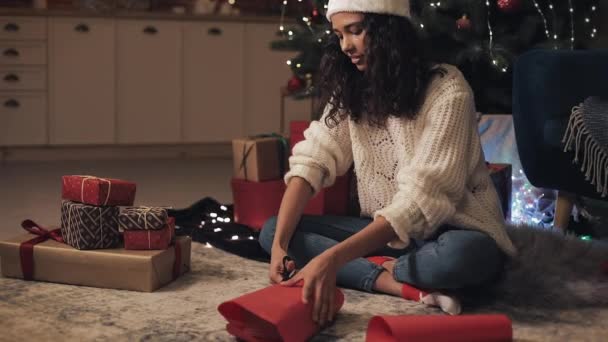 Zooming Pohled na atraktivní Happy Mulatto Girl v Santas klobouku Příprava dárky, Řezací papír, Balení dárky sedí pod vánoční strom na útulném domácím pozadí. Koncept svátkové oslavy. — Stock video