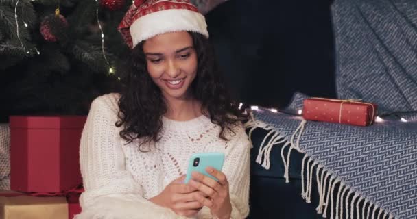 サンタ・ハットの若いプリティガールのクローズアップ休日のクリスマスツリーの下に座ってスマートフォンを入力して使用しながら、装飾されたホーム背景コミュニケーション、休日のコンセプト. — ストック動画