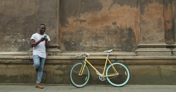 Afroamerikanischer Mann mit Kopfhörer, der sein Smartphone zum Chatten mit Freunden benutzt, während er an eine alte Hauswand gelehnt in der Nähe eines stilvollen modernen Fahrrads steht.. — Stockvideo