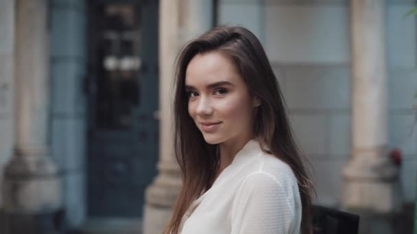 Portret van de jonge charmante Brunette Student Girl met een tas die over haar schouder naar de camera kijkt en glimlacht terwijl ze naar de oude architectuur op de achtergrond staat te kijken. Sluiten. — Stockvideo