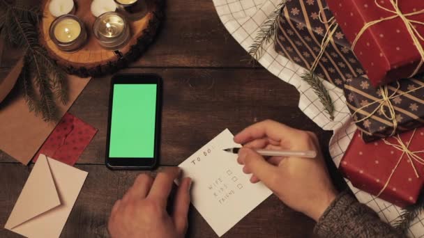 Vista dall'alto delle lancette dei fan Inserire le zecche nell'elenco dei regali per la sua famiglia sulla tavola decorata di Natale in legno con regali avvolti e smartphone con schermo verde su di esso. Chiave cromatica . — Video Stock