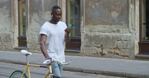 Junger afrikanisch-amerikanischer Mann mit Kopfhörern, der sein Smartphone benutzt, auf den Bildschirm schaut und das stylische moderne Fahrrad schiebt, während er in der Altstadt spaziert. Touristisches Konzept. Seitenansicht. — Stockvideo