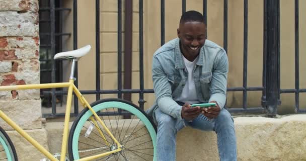 Joven Afroamericano Alegre Usando su Smartphone, Viendo Videos, Mirando Emocionado mientras está sentado cerca de Elegante Bicicleta con Old Metal Fance en el Fondo. Vista frontal . — Vídeos de Stock