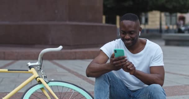 Nahaufnahme eines afrikanisch-amerikanischen Mannes mit Kopfhörern, der sein Smartphone benutzt, den Bildschirm berührt und glücklich und aufgeregt wirkt, während er auf den Stufen neben dem stilvollen Fahrrad im Hintergrund der Stadt sitzt. — Stockvideo