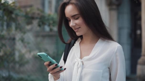 Close Up Shot of Beautiful Student Girl with Bag on her Ramoulder Using her Modern Smartphone, Scrolling the Screen while Standing at Old Building Background. Pomalý pohyb. — Stock video