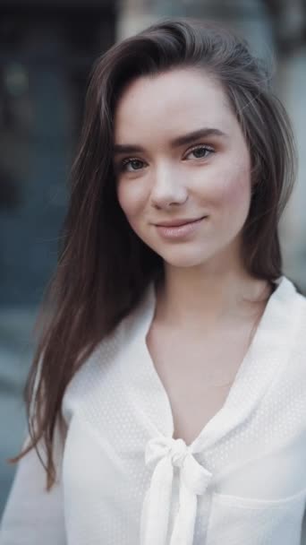 Retrato de la joven morena encantadora girando la cabeza y mirando a la cámara con una sonrisa radiante mientras está de pie al aire libre. Primer plano. Vídeo vertical . — Vídeos de Stock