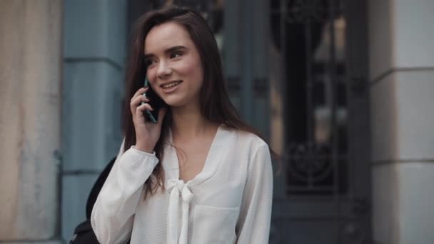 Schöne junge lächelnde brünette Studentin, die auf ihrem Smartphone spricht und im Freien an alter Architektur im Hintergrund spaziert. Kommunikations- und Technologiekonzept. Nahaufnahme. — Stockvideo
