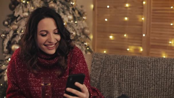 魅力的なブルネットの女性彼女のスマートフォンの画面を見て、笑いながらシャンパンのグラスを保持するコスプレホームで座ってクリスマスツリーの近くの背景. — ストック動画