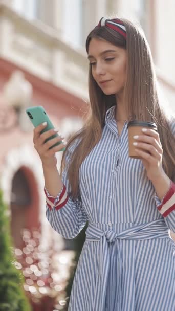 Vídeo Verlical. Mulher encantadora jovem com headband vestindo elegante vestido listrado usando seu smartphone rolagem e sorrindo olhando feliz segurando xícara de café em pé na rua da cidade — Vídeo de Stock