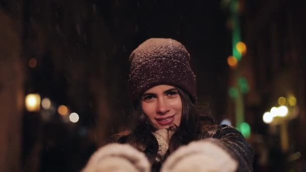 Ritratto di giovane ragazza attraente felice in cappello invernale e guanti che cammina in caduta neve, puntando e sorridendo alla macchina fotografica a sera Street Lights sfondo. Primo piano . — Video Stock