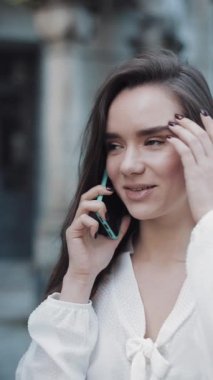 Güzel, gülümseyen esmer kız akıllı telefonuyla konuşuyor arka plandaki eski mimarinin önünde dikiliyor. İletişim ve Teknoloji Konsepti. Dikey Video.