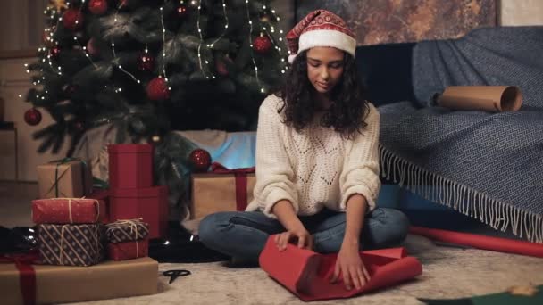 穿着圣诞老人帽和白色套头衫的迷人的快乐女孩准备礼物，包装纸和剪贴纸，坐在圣诞树下，舒适的家庭背景。 假日庆祝概念. — 图库视频影像