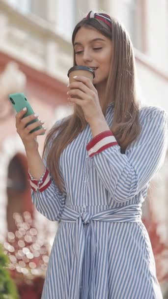 Vídeo Verlical. Joven chica caucásica con encanto con diadema usando elegante vestido a rayas usando su teléfono inteligente Desplazamiento y sonriente mirando feliz beber café de pie en la calle de la ciudad . — Vídeos de Stock