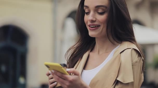 Νεαρή ελκυστική κοπέλα που αναζητούν πραγματικά εντυπωσιασμένος χρησιμοποιώντας το κινητό της τηλέφωνο γέλιο λέγοντας Wow Holding Cofee Κύπελλο Πληκτρολογώντας και στέκεται στο ιστορικό της πόλης από κοντά. — Αρχείο Βίντεο