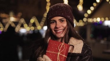 Kış Şapkası Tutma Kutusu Giyen Mutlu Genç Kızın Portresi, Kameraya Bakış ve Gülümseme. Noel Fuarı Işıkları Arkaplanındaki Yağan Kar 'da Duruyor.