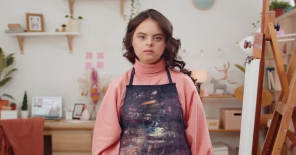 Hübsche Teenagerin mit genetischer Störung, die Friedenszeichen zeigt, während sie vor der Kamera posiert. Porträt eines kleinen talentierten Mädchens in Künstlerschürze, das neben molbert in ihrem Zimmer sitzt. — Stockvideo