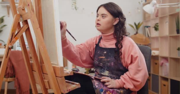 Joven adolescente talentosa que usa pintura de delantal de artistas mientras está sentada frente a Molbert en su habitación. Chica bonita con trastorno genético sosteniendo paleta en sus rodillas creando imagen . — Vídeos de Stock