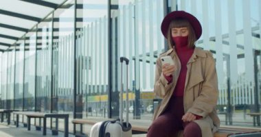 Bayan gezgin bavuluyla bankta oturuyor ve telefon ekranına bakıyor. Pamuk protektif maskeli kadın otobüs durağında otobüs beklerken cep telefonu kullanıyor..